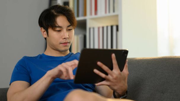 Беззаботный азиатский мужчина фрилансер сидит на диване и проверяет электронную почту или работает онлайн на цифровой планшет. - Фото, изображение