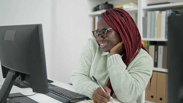 Afrikaanse vrouw met gevlochten haar student met behulp van computer schrijven op notebook in de universiteitsbibliotheek - Foto, afbeelding