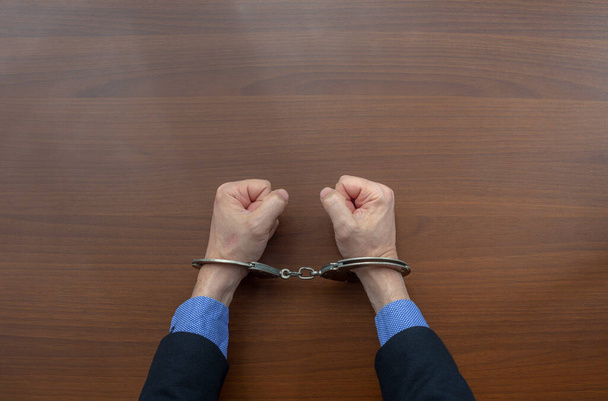 Bovenaanzicht, geboeide handen van een gearresteerd crimineel op de tafel van het politiebureau. kopieerruimte - Foto, afbeelding