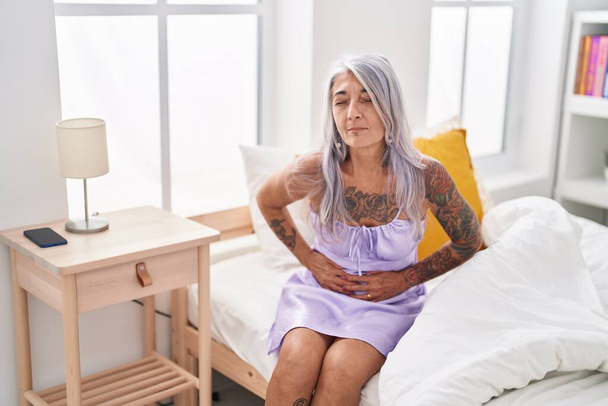 Donna dai capelli grigi di mezza età che soffre di mal di stomaco seduta sul letto in camera da letto - Foto, immagini