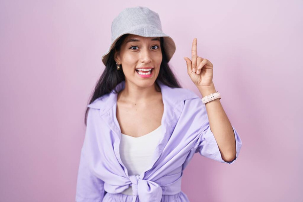 Young hispanic woman standing over pink background wearing hat pointing finger up with successful idea. exited and happy. number one.  - Foto, Bild