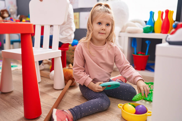 Liebenswertes blondes Mädchen lächelt selbstbewusst auf dem Boden sitzend und spielt im Kindergarten - Foto, Bild