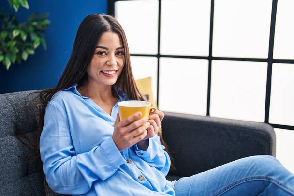 Jovem mulher hispânica bonita bebendo café sentado no sofá em casa - Foto, Imagem