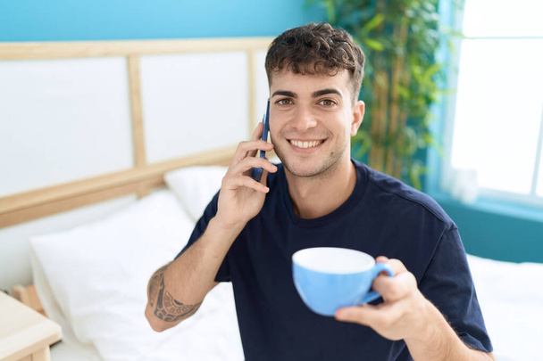 Молодой латиноамериканец разговаривает по смартфону, пьет кофе в спальне - Фото, изображение