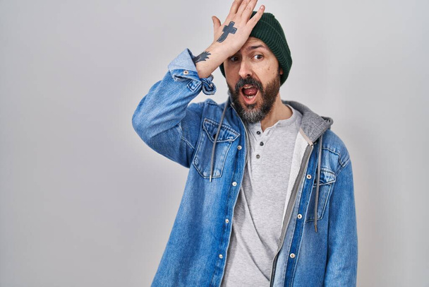 Young hispanic man with tattoos wearing wool cap surprised with hand on head for mistake, remember error. forgot, bad memory concept.  - Foto, afbeelding