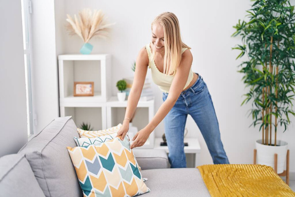 Молодая блондинка улыбается уверенно организуя диван дома - Фото, изображение