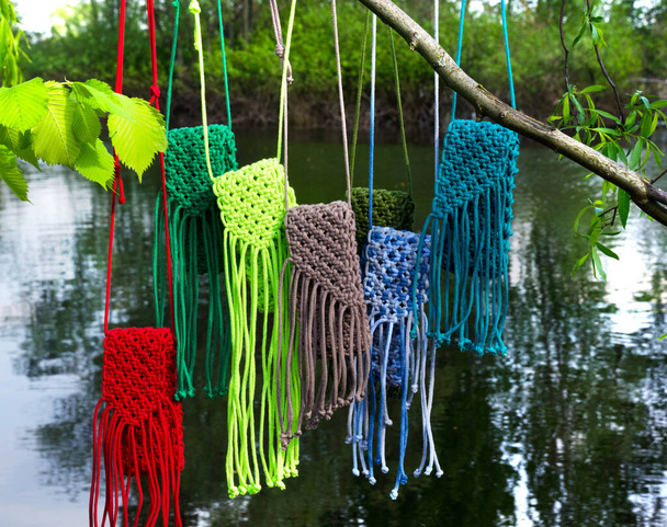 Macrame technikával kötött kis zacskók. Többszínű táskák mobiltelefonhoz a folyó és a zöld fák hátterében. - Fotó, kép