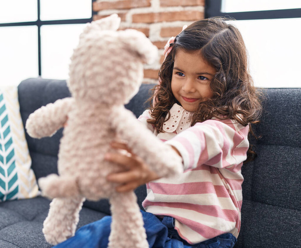 Schattige latino meisje houdt teddy beer zittend op de bank thuis - Foto, afbeelding