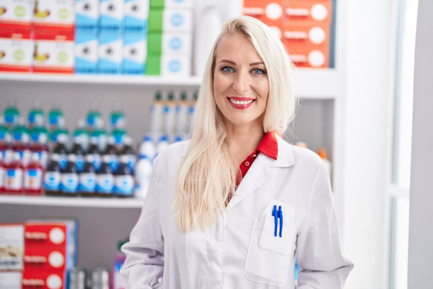 Joven mujer rubia farmacéutica sonriendo confiada de pie en la farmacia - Foto, imagen