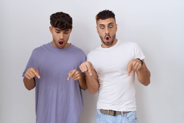 couple gay homosexuel debout sur fond blanc pointant vers le bas avec les doigts montrant la publicité, visage surpris et bouche ouverte  - Photo, image