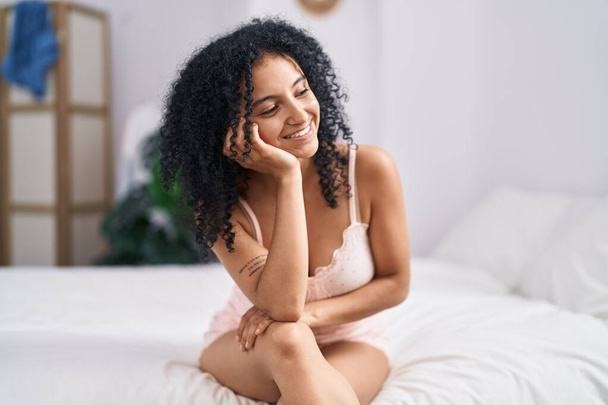 Νεαρή ισπανόφωνη γυναίκα χαμογελά με αυτοπεποίθηση, κάθεται στο κρεβάτι στο υπνοδωμάτιο - Φωτογραφία, εικόνα