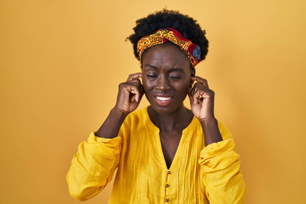 Mujer joven africana con turbante africano cubriendo las orejas con los dedos con expresión molesta por el ruido de la música alta. concepto de sordo.  - Foto, imagen