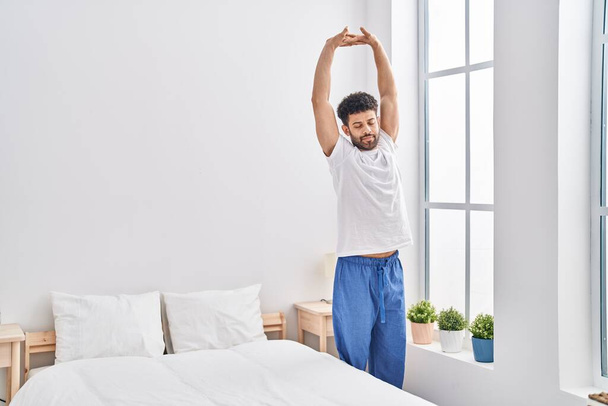 Young arab man waking up stretching arms at bedroom - Фото, изображение