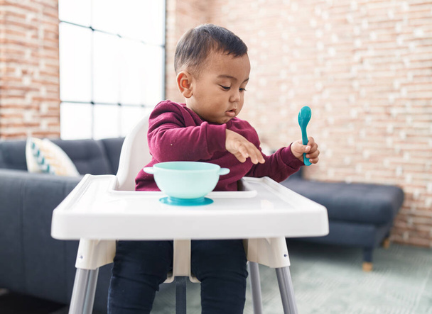 Adorable hispanic boy sitting on highchair holding spoon at home - Photo, image