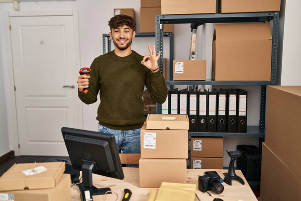 Arabischer Mann mit Bart, der in einem kleinen Geschäft arbeitet und einen Barcode-Scanner hält und mit den Fingern Okay-Zeichen macht, lächelt freundlich und gestikuliert mit einem ausgezeichneten Symbol  - Foto, Bild