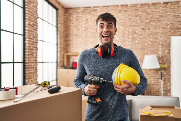 Junger hispanischer Mann, der bei Renovierungsarbeiten zu Hause arbeitet, feiert verrückt und staunt über den Erfolg mit offenen Augen, die aufgeregt schreien.  - Foto, Bild