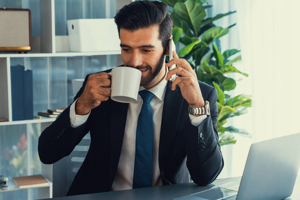Businessman working in modern office workspace with cup of coffee in his hand while answering phone call making sales calls or managing employee. Fervent - Фото, изображение