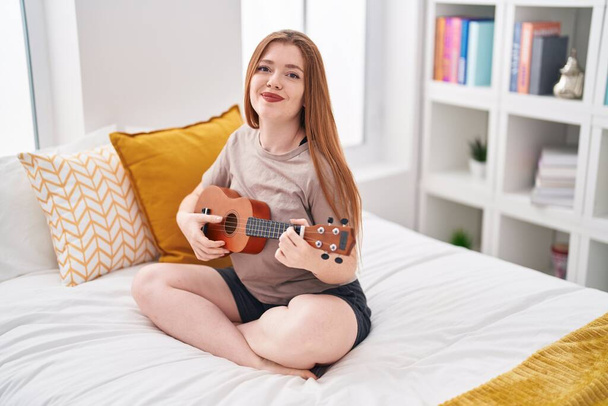 Young redhead woman playing ukulele sitting on bed at bedroom - Photo, Image