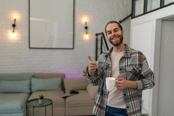Handsome positive man holding cup of fresh coffee or tea in hands enjoying free time with hot beverage, showing thumb up, like sign. - Foto, Bild