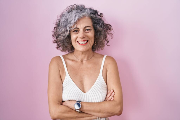 Middle age woman with grey hair standing over pink background happy face smiling with crossed arms looking at the camera. positive person.  - Fotografie, Obrázek