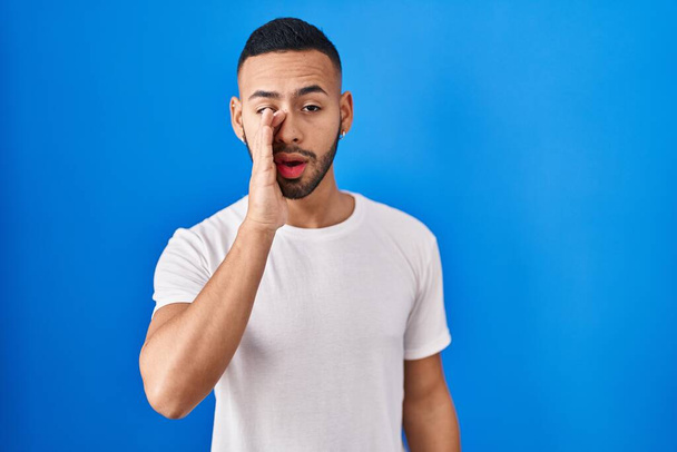 Young hispanic man standing over blue background hand on mouth telling secret rumor, whispering malicious talk conversation  - Fotografie, Obrázek