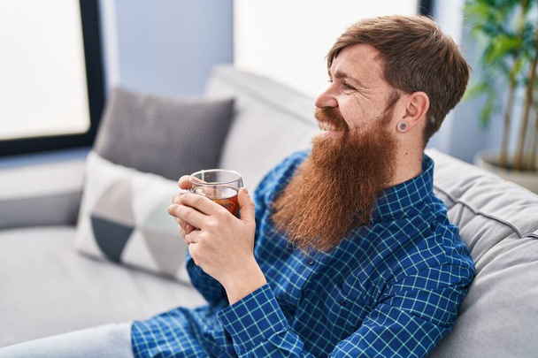 Young redhead man drinking tea sitting on sofa at home - Foto, immagini
