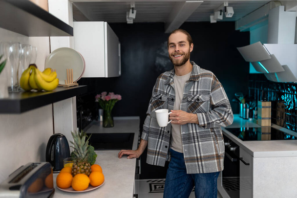 Χαμογελαστή νεαρός άνδρας με γενειάδα κρατώντας κύπελλο με καφέ, στέκεται δίπλα στον πάγκο της κουζίνας με σύγχρονο φως εσωτερικό, απολαμβάνοντας φρέσκο ζεστό ποτό στο σπίτι. - Φωτογραφία, εικόνα