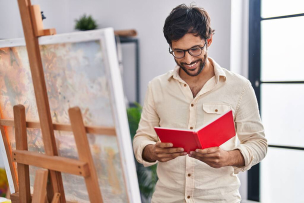 Młody Latynos mężczyzna artysta uśmiecha się pewnie czytając książkę w studio sztuki - Zdjęcie, obraz