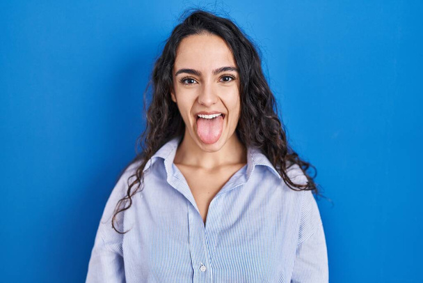 Young brunette woman standing over blue background sticking tongue out happy with funny expression. emotion concept.  - Foto, Imagem