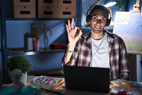 Молодой латиноамериканец, сидящий в арт-студии с ноутбуком поздно ночью, улыбаясь позитивно, делает хорошо знак с рукой и пальцами. успешное выражение.  - Фото, изображение