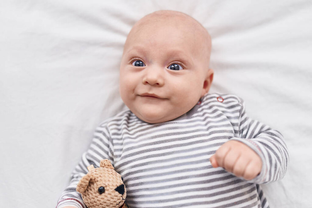 Sevimli beyaz bebek, yatak odasında oyuncak ayıyla yatakta kendinden emin şekilde gülümsüyor. - Fotoğraf, Görsel