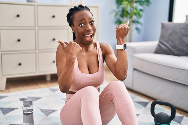 Piękna czarna kobieta za pomocą inteligentnego zegarka treningowego w domu wskazując kciuk do góry na stronę uśmiechnięta szczęśliwa z otwartymi ustami  - Zdjęcie, obraz