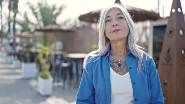 Keski-ikäinen harmaa tukka nainen hymyilee luottavainen seisoo kädet ristissä ele kahvilan terassilla - Materiaali, video