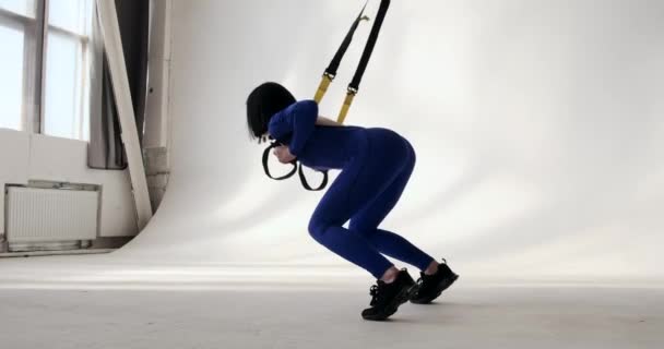 Fit žena v modrém atletickém oblečení předvádí dřepy, zatímco se naklání dopředu a pomocí odporu pásy pro zvýšení odporu. Celotělové cvičení pro tónování a posilování svalů. - Záběry, video
