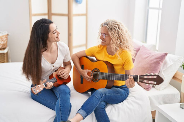 Two women mother and daughter playing classical guitar and ukulele at bedroom - Fotó, kép