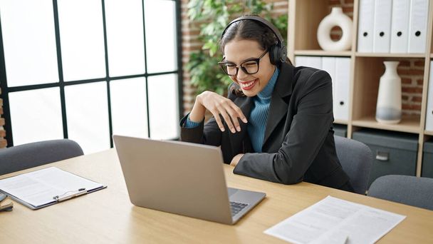 Νεαρή όμορφη ισπανίδα επιχειρηματίας χρησιμοποιώντας φορητό υπολογιστή φορώντας ακουστικά στο γραφείο - Φωτογραφία, εικόνα