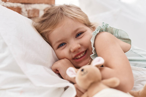 Αξιολάτρευτο ξανθό κορίτσι κρατώντας κούκλα ξαπλωμένη στο κρεβάτι στο υπνοδωμάτιο - Φωτογραφία, εικόνα