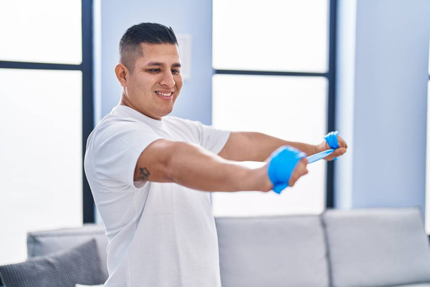 Jovem latino sorrindo confiante usando treinamento elástico em casa - Foto, Imagem
