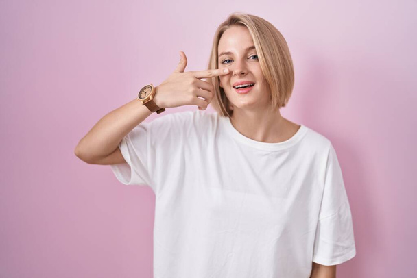Young caucasian woman standing over pink background pointing with hand finger to face and nose, smiling cheerful. beauty concept  - Zdjęcie, obraz