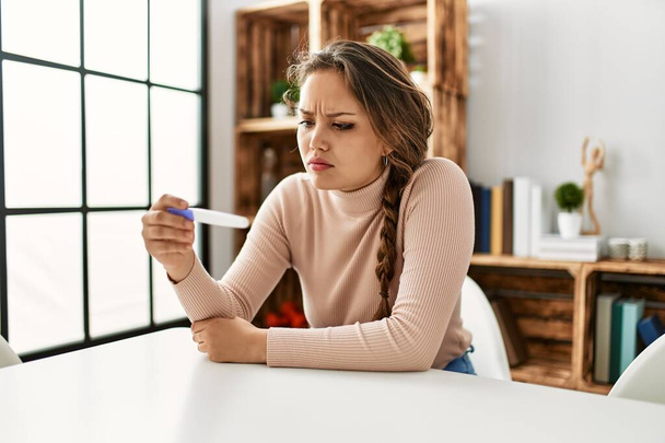 Young beautiful hispanic woman holding pregnancy test sitting on table at home - Photo, image