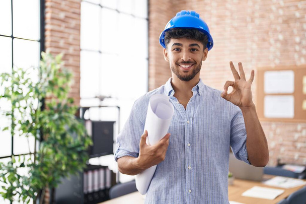 Homme arabe avec barbe portant un casque d'architecte au bureau de construction faire ok signe avec les doigts, sourire geste amical excellent symbole  - Photo, image