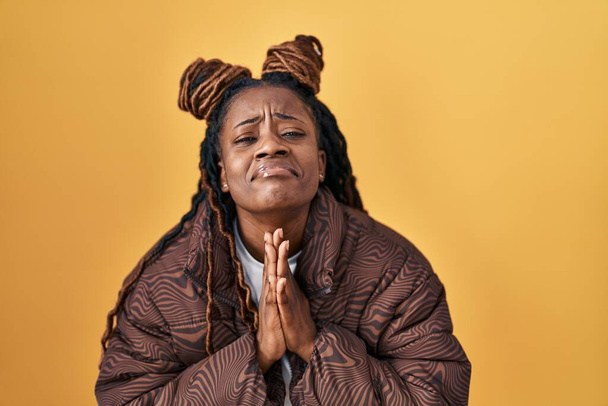 Африканська жінка з плетеним волоссям стоїть над жовтим фоном, благаючи і молячись руками разом з надією на обличчя дуже емоційно і хвилюючо. благання..  - Фото, зображення