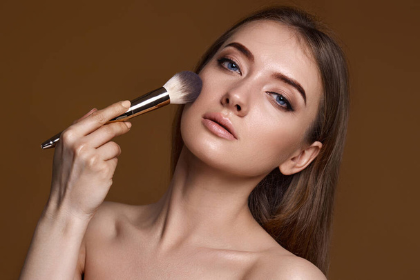 young woman applying blush on cheeks with make-up brush on beige background - Foto, Imagem