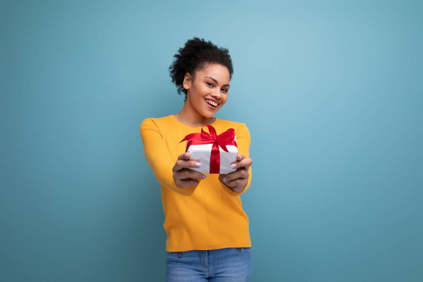 szczęśliwa młoda Latynoska kobieta z czarnymi kręconymi włosami w żółtym swetrze trzyma pudełko prezentów. - Zdjęcie, obraz