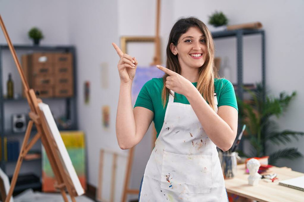 Giovane donna bruna in studio d'arte sorridente e guardando la fotocamera che punta con due mani e le dita di lato.  - Foto, immagini