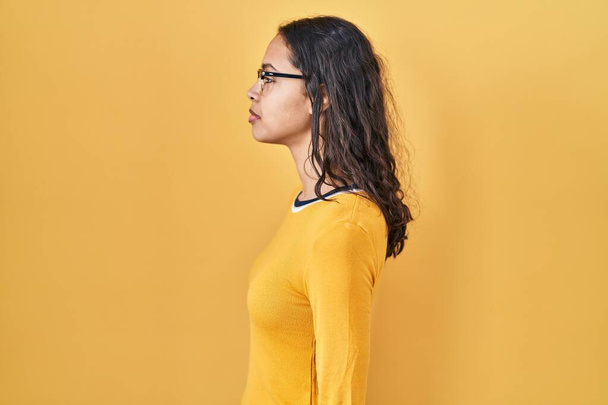 Молодая бразильская женщина в очках на желтом фоне глядя в сторону, расслабиться профиль позировать с естественным лицом с уверенной улыбкой.  - Фото, изображение