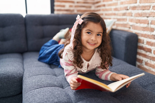 Adorable chica hispana leyendo libro tumbado en el sofá en casa - Foto, Imagen