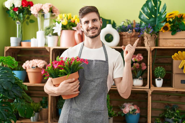 Hispanic man with beard working at florist shop holding plant smiling happy pointing with hand and finger to the side  - Fotoğraf, Görsel
