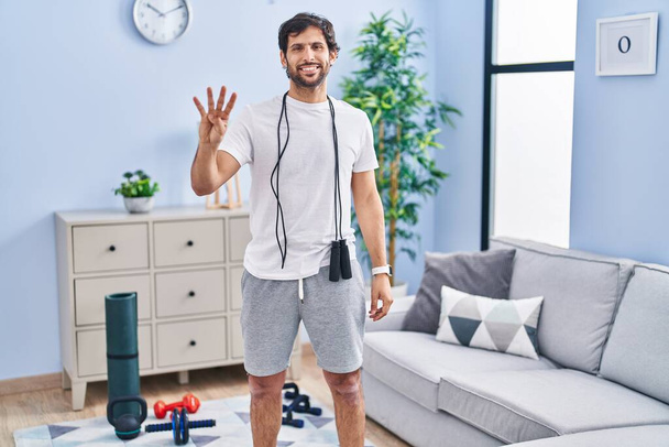 Hombre latino guapo usando ropa deportiva en casa mostrando y apuntando hacia arriba con los dedos número cuatro mientras sonríe confiado y feliz.  - Foto, imagen