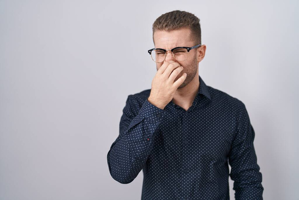 Young caucasian man standing over isolated background smelling something stinky and disgusting, intolerable smell, holding breath with fingers on nose. bad smell  - Фото, зображення